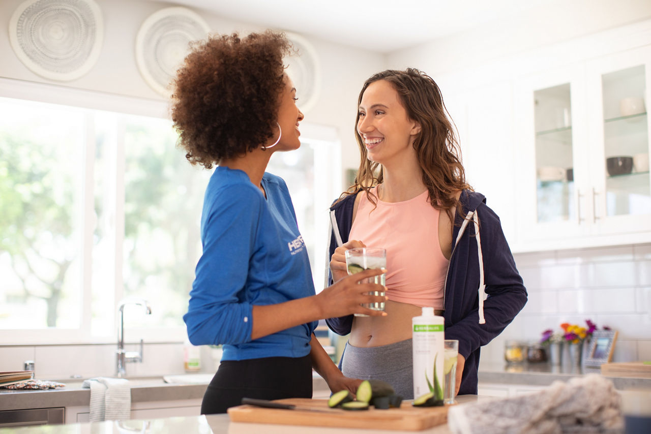 Two women smiling and sharing an aloe drink in their kitchen