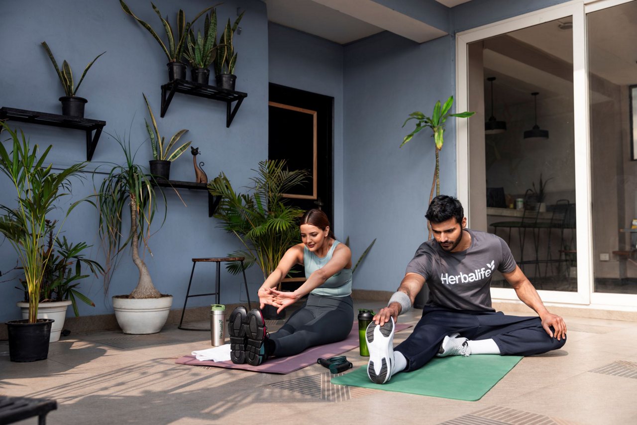 एक जोड़ा घर में योग मैट पर योग अभ्यास कर रहे हैं, और पास में Herbalife की बोतल रखी हुई है।