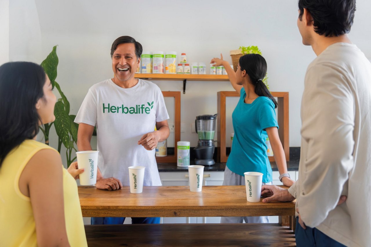 लोगों का एक समूह हर्बालाइफ कप के साथ रसोई काउंटर के चारों ओर इकट्ठा हुआ, जहां एक सफेद हर्बालाइफ टी-शर्ट पहने एक व्यक्ति अन्य लोगों के साथ बातचीत कर रहा था, जबकि शेल्फ पर उत्पाद के कंटेनर दिखाई दे रहे थे।