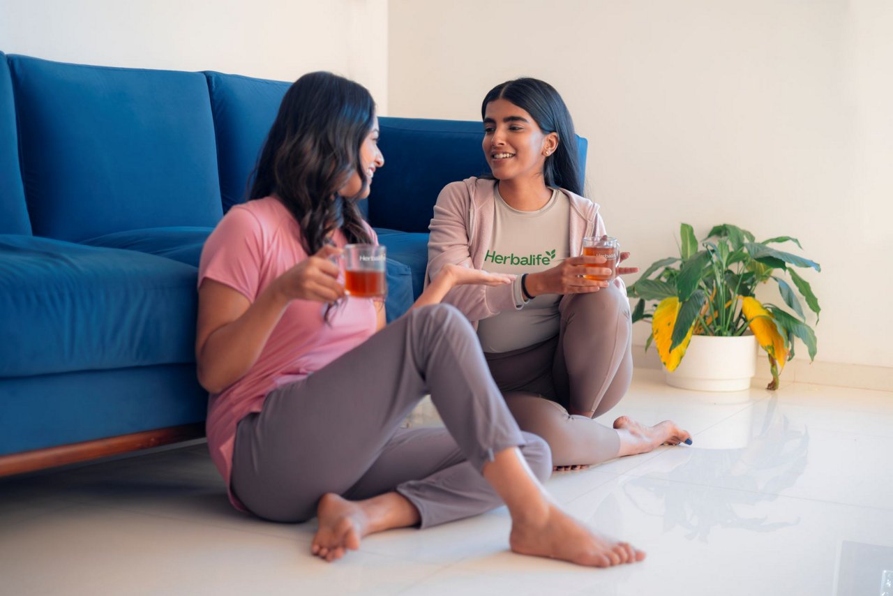 Two women sitting on floor by blue couch enjoying Herbalife tea in glass cups, one wearing Herbalife t-shirt, engaged in conversation with potted plant nearby.