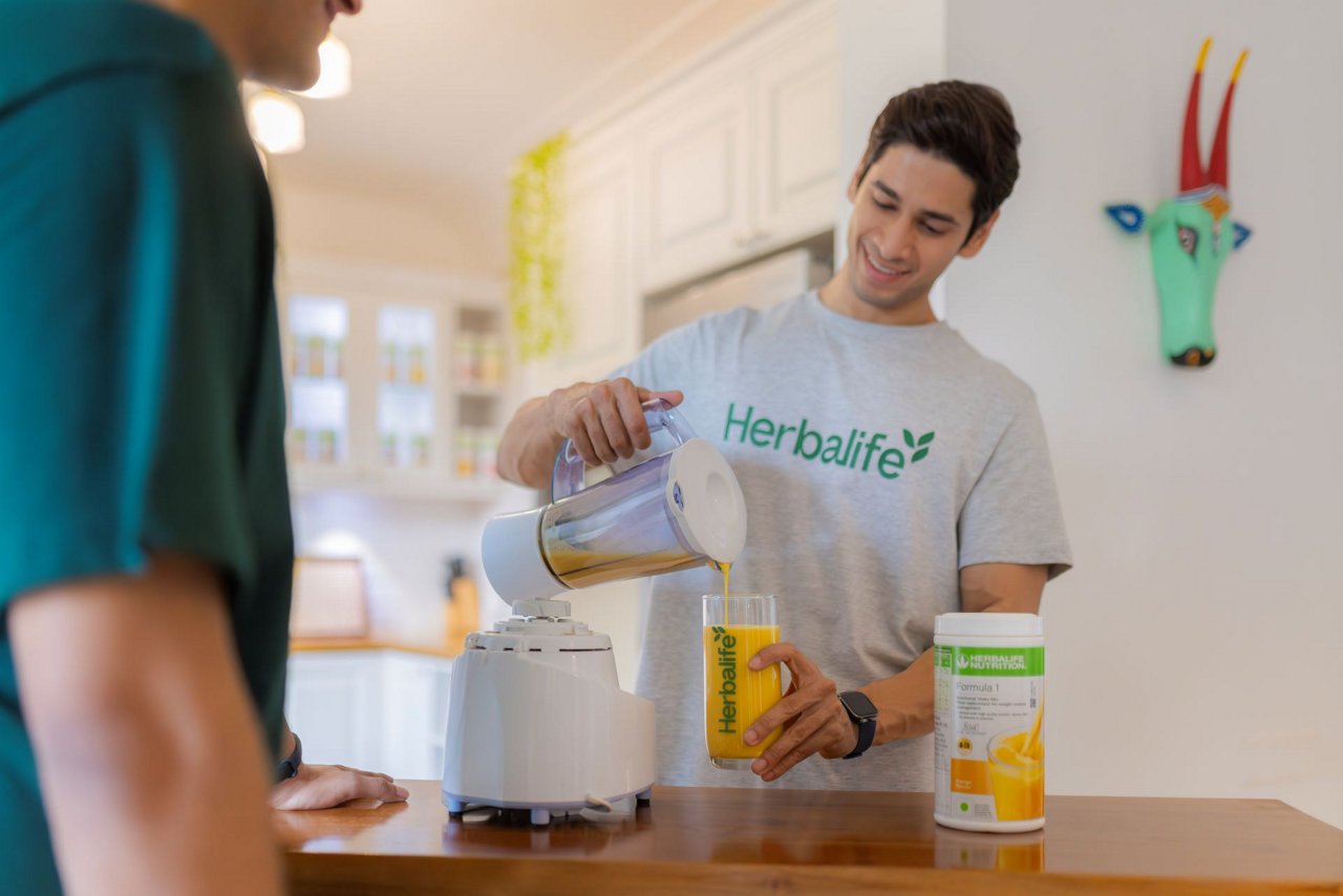 Herbalife टी-शर्ट पहने हुए और फॉर्मूला 1 शेक डालता हुए एक व्यक्ति।