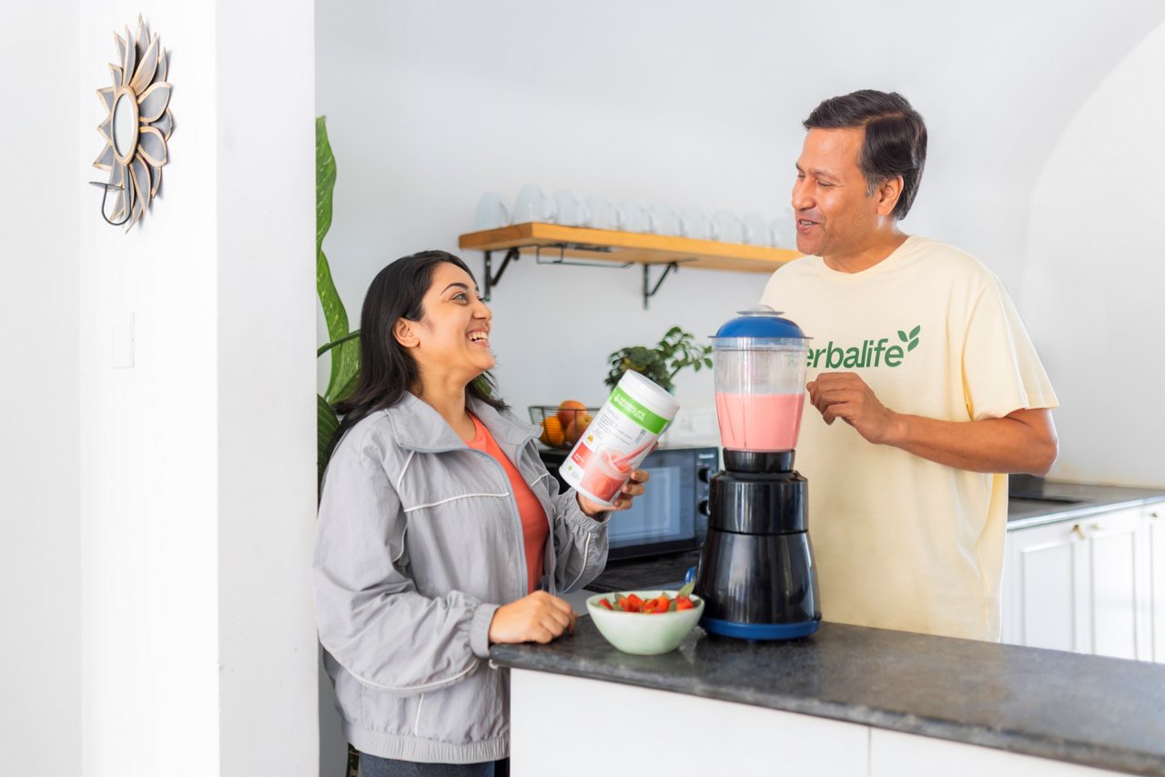 एक पुरुष और एक महिला आधुनिक रसोई में हर्बालाइफ स्ट्रॉबेरी-स्वाद वाले पाउडर का उपयोग करके स्ट्रॉबेरी शेक तैयार कर रहे हैं