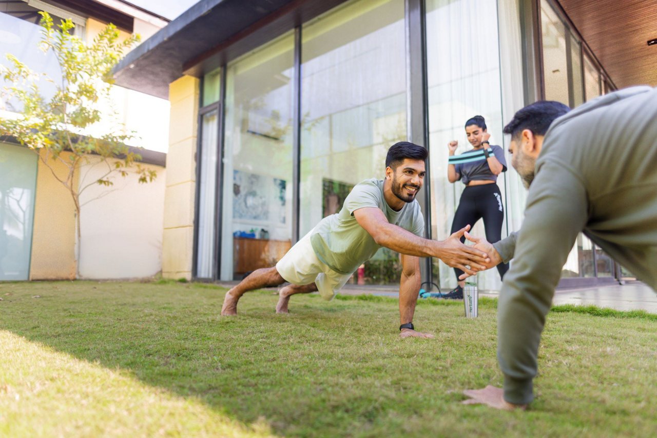 दो पुरुष पिछवाड़े में घास पर पुशअप्स कर रहे हैं, जबकि एक महिला कांच की दीवारों वाले एक आधुनिक घर के आंगन में काम कर रही है।