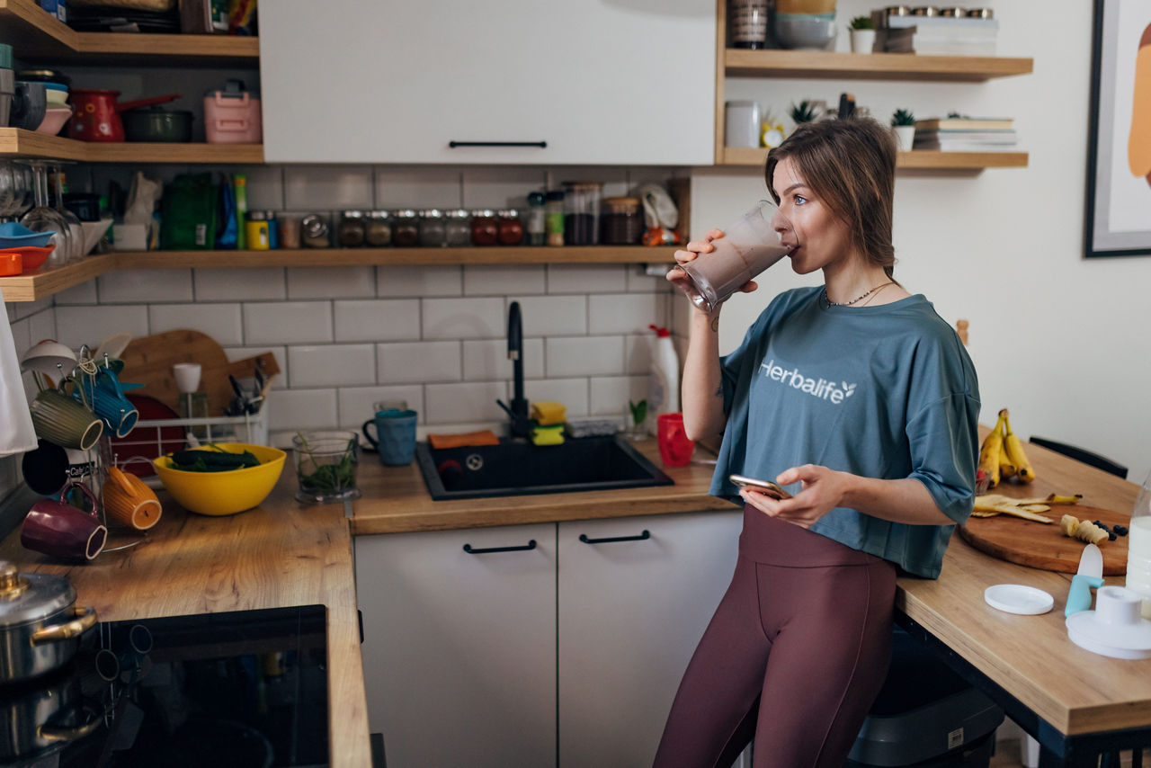 Woman drinking a shake in the kitchen with her phone in her hand