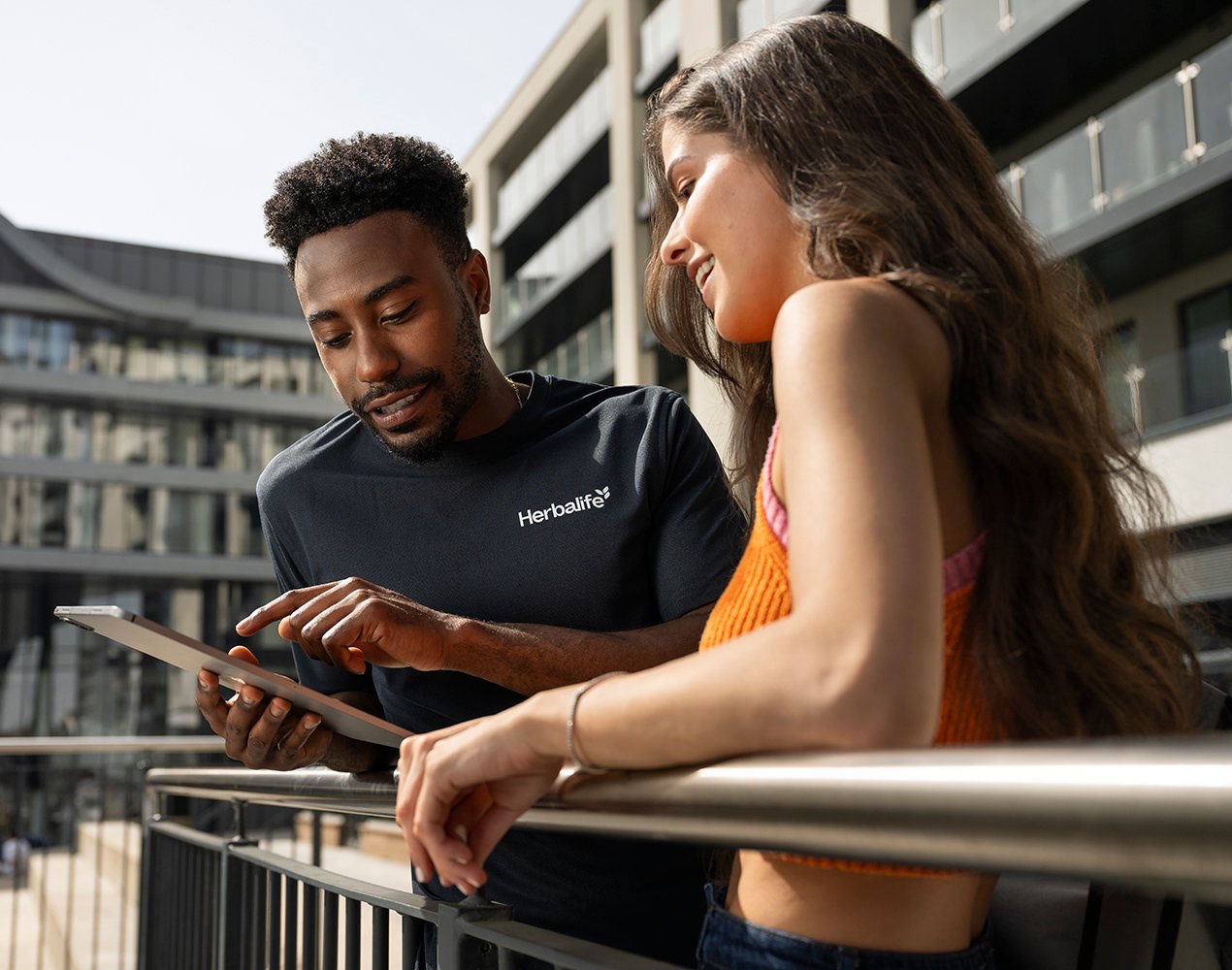 Two adults stand on an outdoor balcony, one holding a tablet while the other points at the screen, with modern buildings in the background.