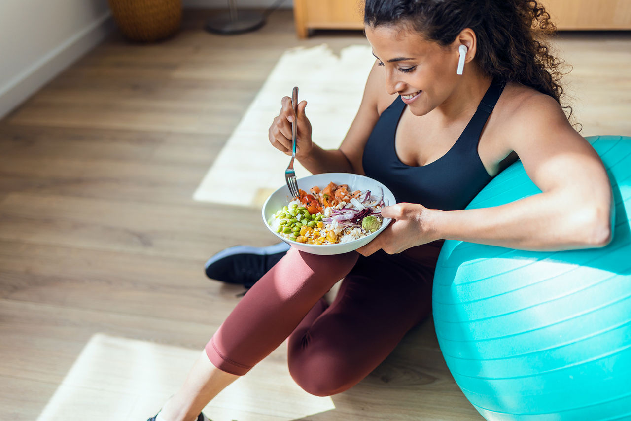 Shot of sporty young woman eating healthy while listening to music sitting on the floor at home.