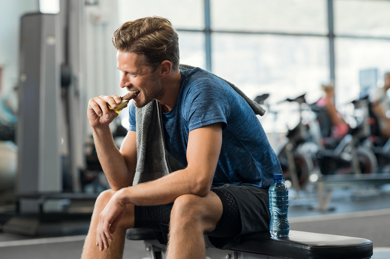 Sweaty young man eating energy bar at gym. Handsome mid guy enjoying chocolate after a heavy workout in fitness studio. Fit man biting a snack and resting on bench.