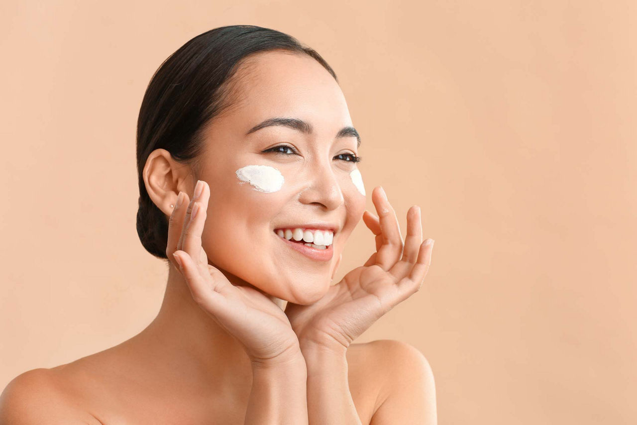Beautiful Asian woman applying cream to her face against a color background