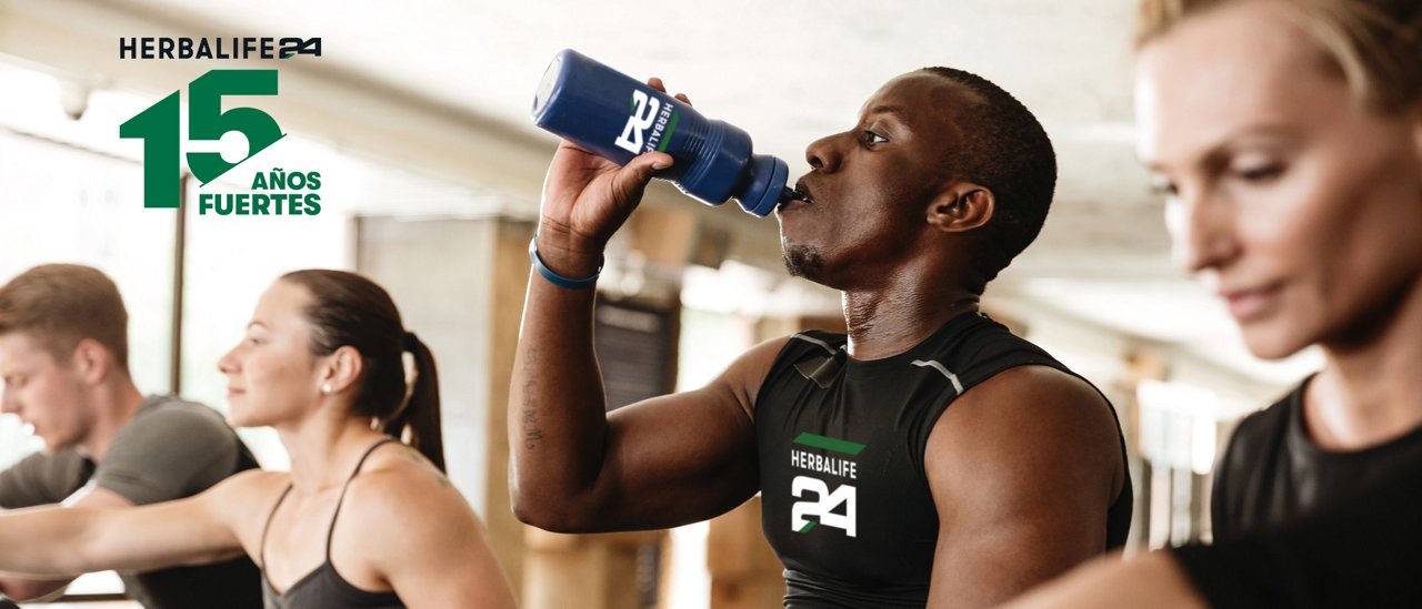 Personas utilizando equipos de ejercicio en un gimnasio; una persona bebe de una botella deportiva mientras otras entrenan al fondo.