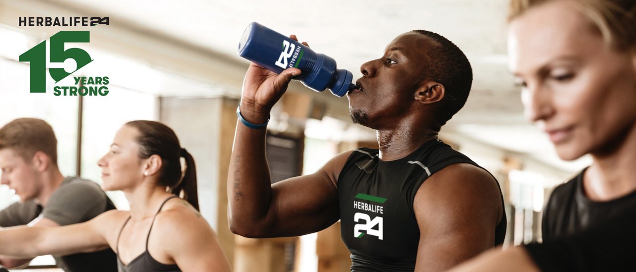 People using exercise equipment in a gym; one person drinks from a sports bottle while others exercise in the background.