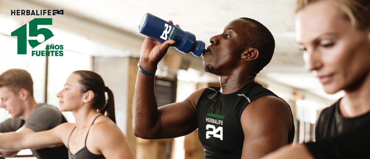 Personas utilizando equipos de ejercicio en un gimnasio; una persona bebe de una botella deportiva mientras otras entrenan al fondo.