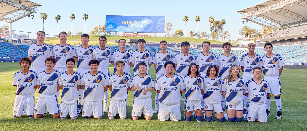 LA Galaxy Special Olympics Unified Team players stand together in full uniform for a group photo on the field at Dignity Health Sports Park, with the stadium seating and scoreboard visible in the background.