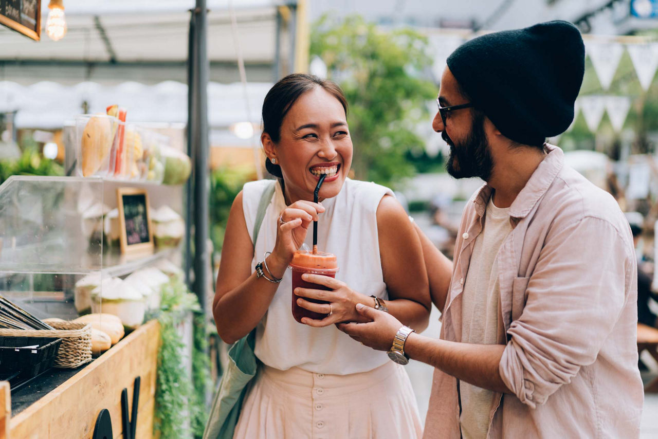 Girlfriend drinking a smoothie drink in front of a vendor street shop, while her boyfriend is standing next to her. Both of them are smiling and looking at each other.
