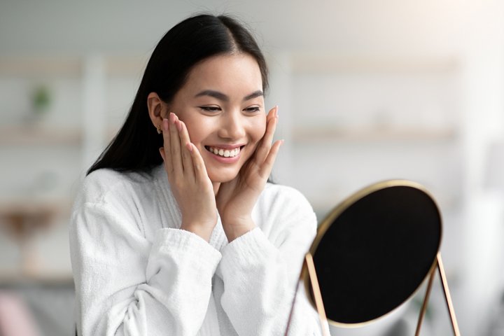 A woman looking in the mirror with both hands holding the soft skin of her cheeks