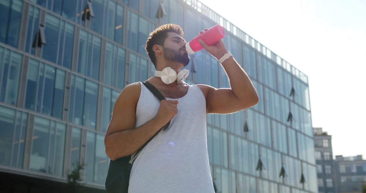Tired young man runner drinking water, relaxing after sport training outdoors. Indian male drink water walking from gym outdoors in city