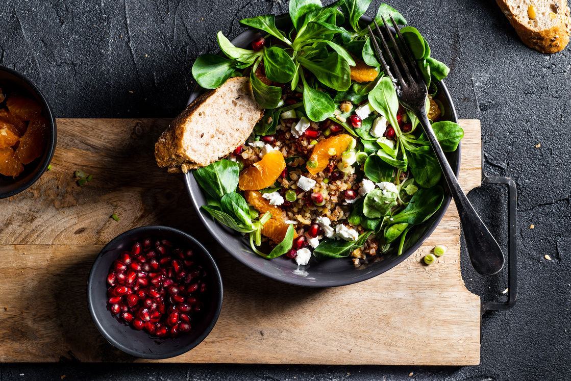 Buckwheat salad with lamb's lettuce, pomegranat seeds, goat cheese, mandarine and spring onion, Served with whole grain baguette and red wine. Black table and black background.