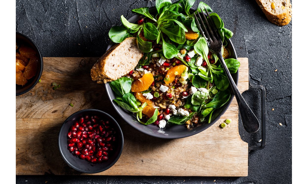 Buckwheat salad with lamb's lettuce, pomegranat seeds, goat cheese, mandarine and spring onion, Served with whole grain baguette and red wine. Black table and black background.