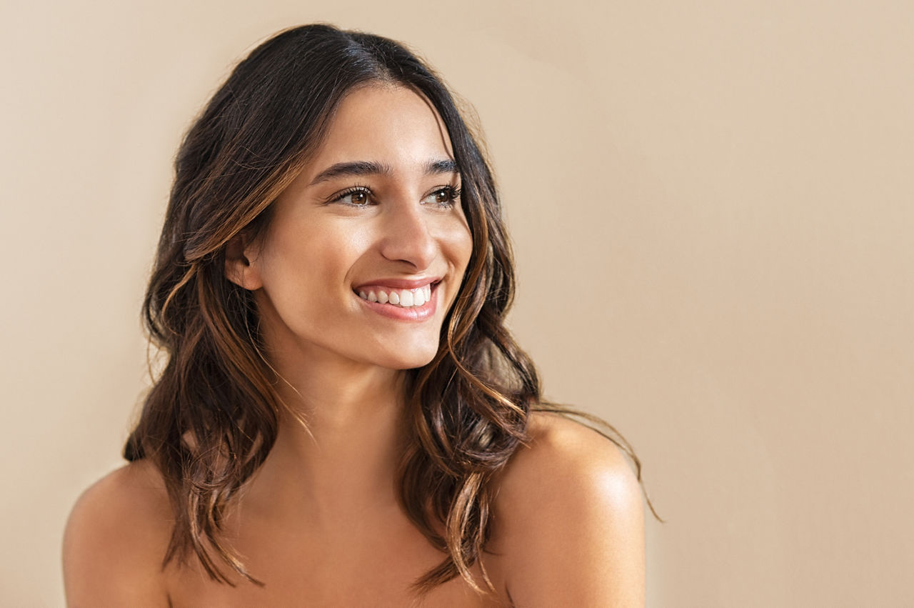 Happy beauty woman looking away isolated on background. Cheerful young brunette with nude shoulder smiling after skin treatment. Beautiful hispanic girl waiting spa therapy.