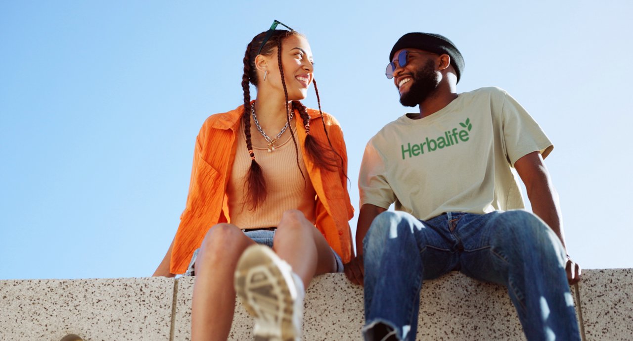 Dwoje uśmiechniętych młodych ludzi siedzi na murku pod błękitnym niebem; mężczyzna ma na sobie koszulkę z logo Herbalife.