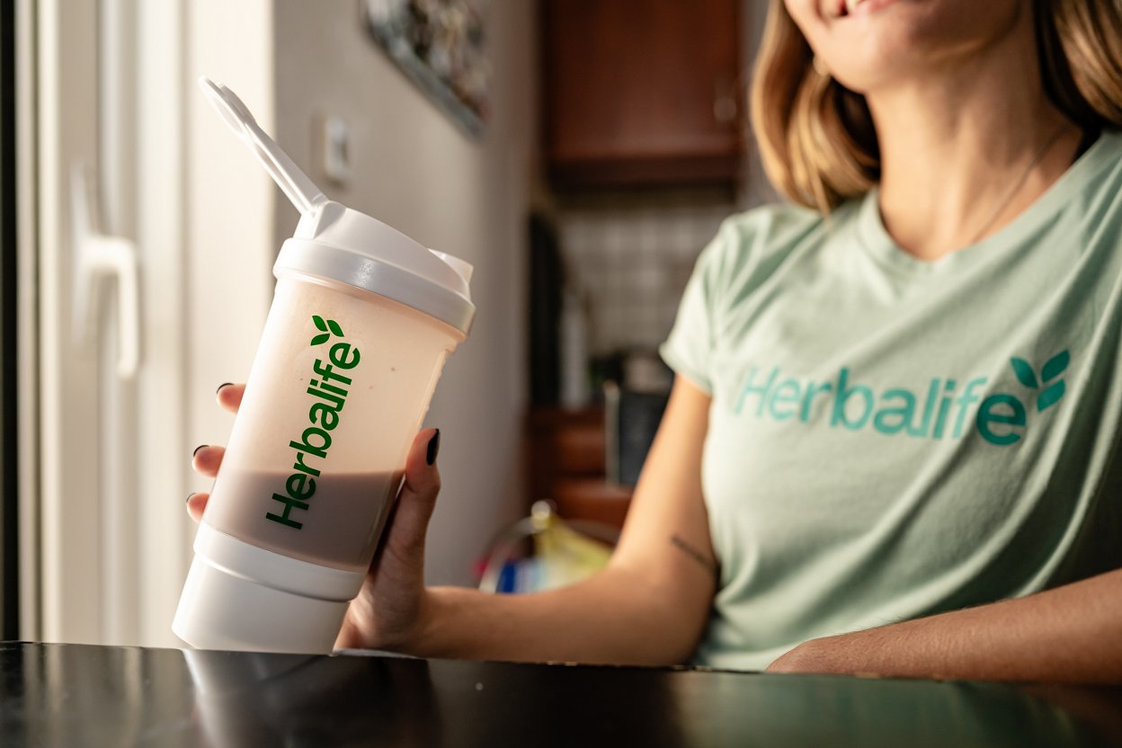 Woman with a shake wearing a green Herbalife-branded t-shirt in modern kitchen.