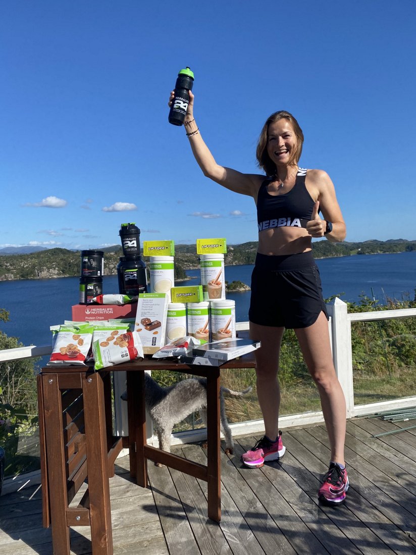 Et bilde av Annette Velde Sande, Herbalife-sponset idrettsutøver, ved siden av et bord fylt med Herbalife-produkter og med en Herbalife24® sportsflaske i hånden.