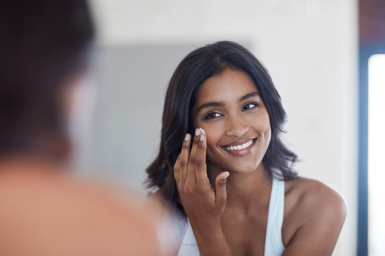 Skincare, woman and face and reflection in bathroom mirror for grooming and morning routine at home. Indian, female person and dermatology for natural shine or glow with collagen lotion and cream.