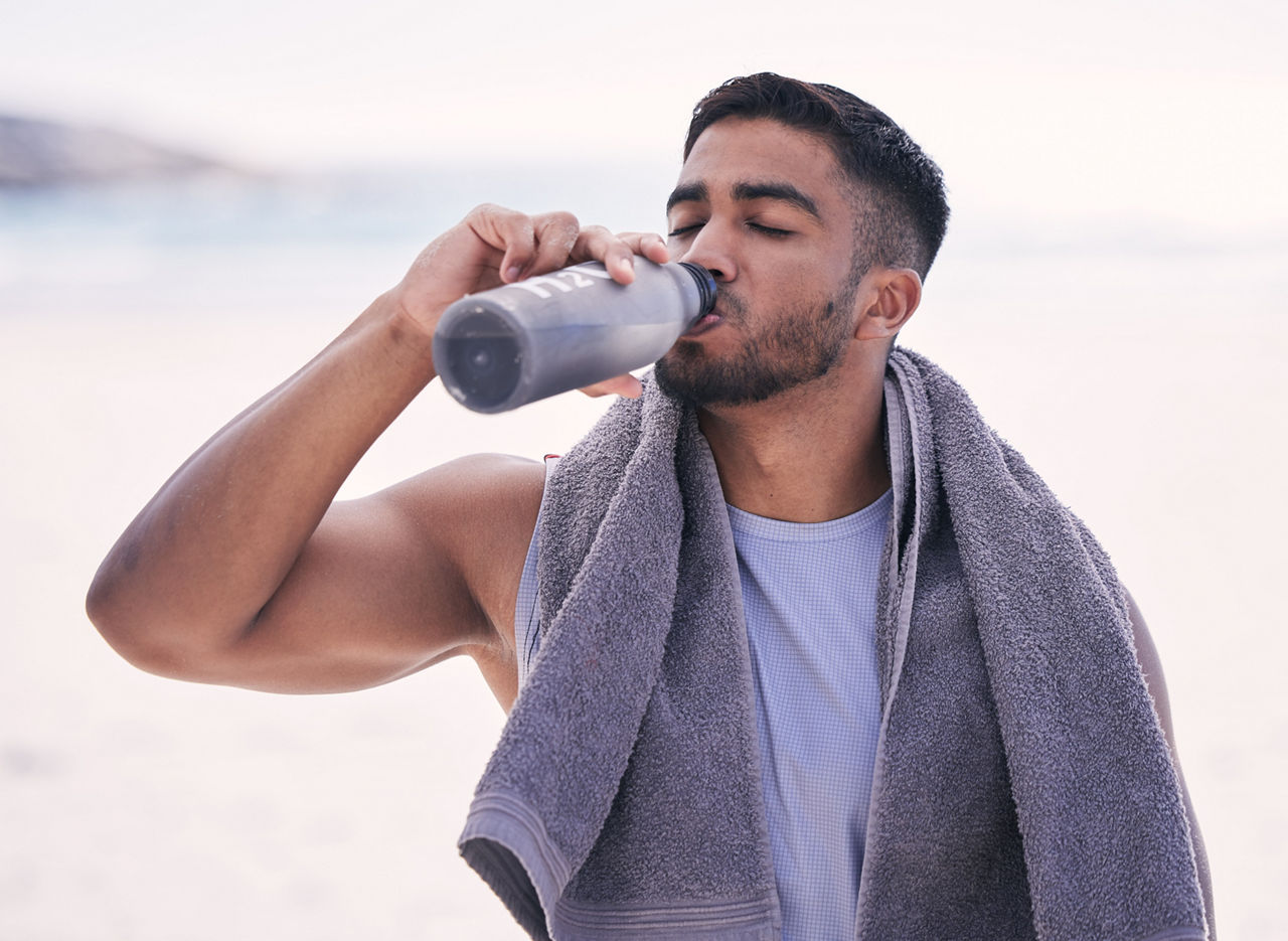 Fitness, sports and man drinking water at the beach after running, training or morning cardio workout with bottle. Exercise, break and thirsty Indian male runner or person with hydration drink at sea.