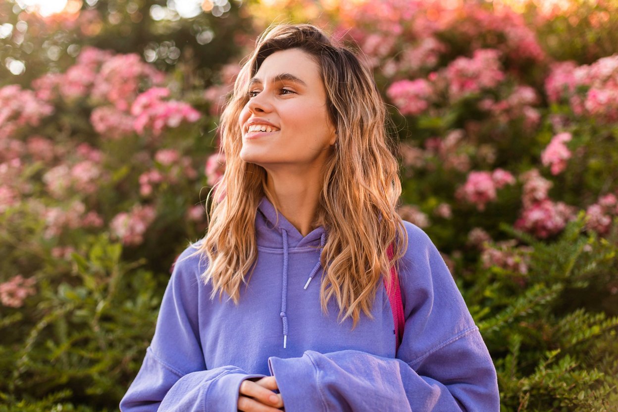 Smiling girl puts on a hood with red hoodie. Pretty young caucasian woman averts her gaze posing on pink background with flowers. Curly blonde woman wear purple hoody. Mood, lifestyle, concept.